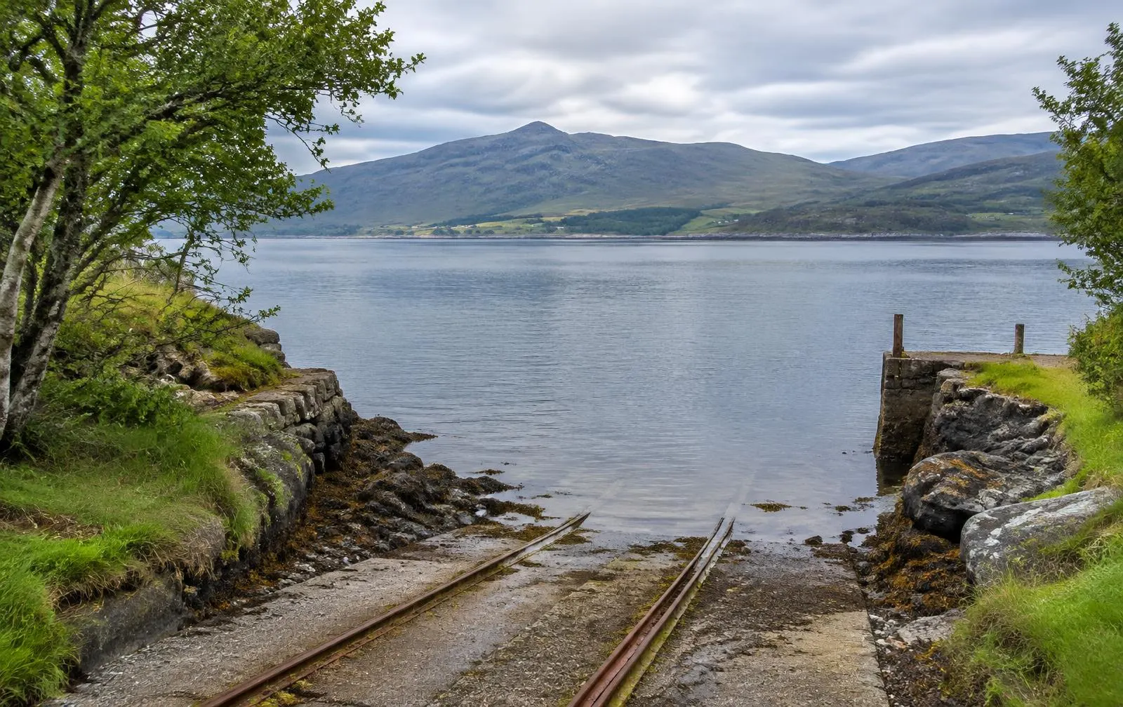 A ferry slipway on a wooded island shore looking across a calm sound to mainland hills